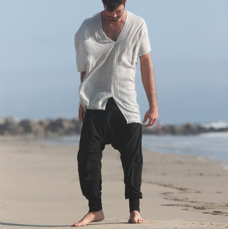 May include: A man wearing a white, loose-fitting shirt with a V-neck and black, loose-fitting pants. He is standing on a sandy beach.