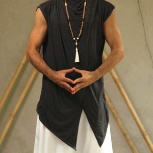Può includere: Una tunica senza maniche grigio scuro con orlo asimmetrico, abbinata a pantaloni bianchi. Una lunga collana di perline con nappa pende dallo scollo. L'outfit ha uno stile rilassato e fluido, adatto per yoga o meditazione.