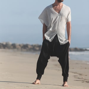 May include: A person wearing a white, loose-fitting shirt with a v-neck and a silver necklace. They are wearing black pants with a loose fit and are standing on a sandy beach.
