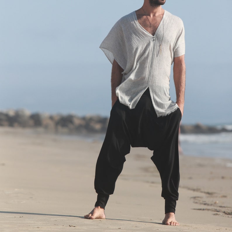 May include: A person wearing a white, loose-fitting shirt with a v-neck and a silver necklace. They are wearing black pants with a loose fit and are standing on a sandy beach.