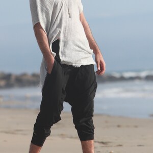 May include: A person wearing a white, loose-fitting top and black, cropped pants. The pants have a unique, gathered design at the ankles.