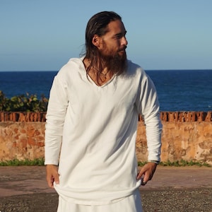 Könnte beinhalten: Ein weißes Langarmshirt mit V-Ausschnitt. Dazu wird ein passender weißer Rock getragen. Der Hintergrund zeigt ein blaues Meer und einen blauen Himmel, mit einer Steinmauer im Vordergrund. Das Outfit ist schlicht und elegant.