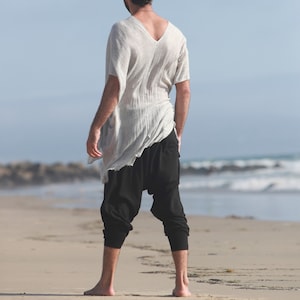 May include: A person wearing a white, loose-fitting, long-sleeved shirt with a V-neck and black, loose-fitting pants. The person is standing on a sandy beach.