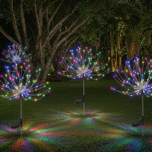 May include: Three outdoor solar garden lights shaped like dandelion spheres, each featuring thin, flexible wire stems radiating outward with small, multicolored LED lights that cast a vibrant, starburst-like glow onto the grass below.