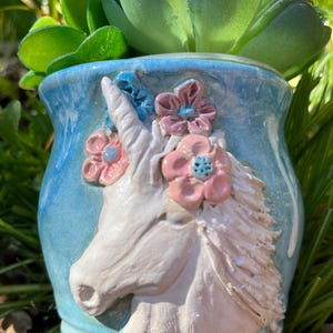 May include: A light blue ceramic planter featuring a white unicorn design and pink and blue floral accents. The planter is filled with a green succulent plant. The planter is approximately 10 cm tall.