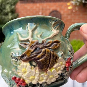 May include: A ceramic mug with a glossy green glaze featuring a raised, three-dimensional relief of a brown deer head with antlers, surrounded by white daisy flowers and clusters of small red berries on the front surface of the vessel.