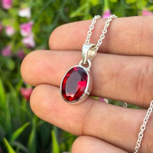 Pode incluir: Um colar com pendente em prata apresentando uma pedra preciosa de cor vermelho-escura, de formato oval facetado, engastada numa moldura de prata polida. O pendente pende de uma corrente de prata delicada, realçando a clareza e a cor viva.