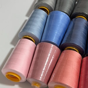 Könnte beinhalten: Eine Sammlung farbiger Garnrollen in verschiedenen Rosa-, Blau-, Grau- und Weißtönen. Die Rollen sind in Klarsichtfolie verpackt und in Reihen angeordnet, die eine Reihe von Farben für Näh- und Bastelprojekte zeigen.