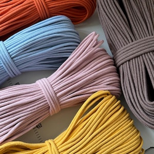 May include: A collection of colorful macrame cords in various hues. The image showcases bundles of orange, light blue, pink, yellow, and brown cords. Each cord is neatly coiled and bound, highlighting the texture and color variations. Ideal for crafting and DIY projects.