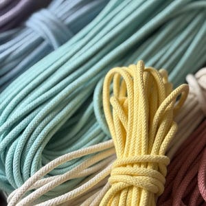 Puede incluir: Primer plano de varios haces de cordón de macramé colorido en tonos lavanda, azul, amarillo, crema y rosa. Los cordones están bien enrollados y listos para proyectos de manualidades. La imagen destaca la textura y la variedad de colores.