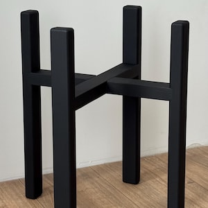May include: A black wooden plant stand. It has a square top and four rectangular legs, joined by a cross-shaped support. The stand is designed for a plant pot and is suitable for indoor or outdoor use. The stand's height is approximately 40 cm.