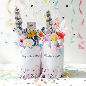 Può includere: Due contenitori di carta bianchi con bordi smerlati, con disegni floreali e le frasi "Happy Birthday" e "Alles Gute zum" in scrittura nera. I contenitori sono riempiti di fiori e verde di carta colorata. Nastri e coriandoli completano la scena festosa.