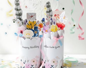 May include: Two white, scalloped-edge paper containers with floral designs and the phrases "Happy Birthday" and "Alles Gute zum" in black script. The containers are filled with colorful paper flowers and greenery. Ribbons and confetti add to the festive scene.