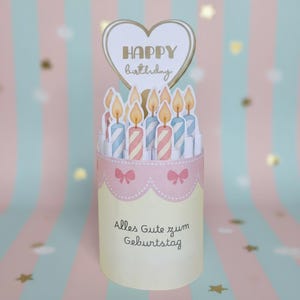 Könnte beinhalten: Eine Pop-up-Geburtstagskarte in Kuchenform mit Kerzen mit blau-rosa Streifen. Ein herzförmiger Aufsatz mit der Aufschrift "HAPPY birthday". Der Kuchen hat eine rosa Borte mit Schleifen und dem Text "Alles Gute zum Geburtstag".