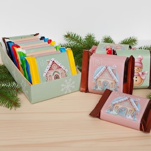 Puede incluir: Un calendario de adviento con golosinas numeradas en una caja verde claro. Las golosinas están envueltas en papel rosa y verde, con ilustraciones de casas de jengibre y un muñeco de nieve. La caja está decorada con copos de nieve.
