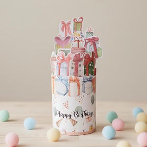 May include: A cylindrical birthday card featuring watercolor illustrations of wrapped gifts with bows. The card has a pop-up design with gifts stacked on top. The words "Happy Birthday" are written on the front of the card.