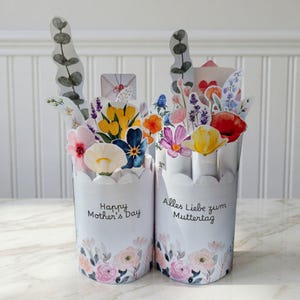 May include: Two white paper flower pots with scalloped edges, each filled with colorful paper flowers. One pot says "Happy Mother's Day," the other says "Alles Liebe zum Muttertag." Floral designs adorn the base of the pots.
