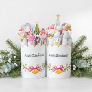 Könnte beinhalten: Zwei weiße Adventskalender-Behälter mit gewellten Rändern, verziert mit festlichen Illustrationen und dem Text "Adventskalender". Jeder Behälter hat eine Schleife, Glocken und Grünzeug. Die Behälter sind mit nummerierten, weihnachtlichen Ausschnitten gefüllt.