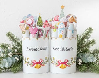 Op de afbeelding: Twee witte adventskalendercontainers met geschulpte randen, versierd met feestelijke illustraties en de tekst "Adventskalender". Elke container heeft een strik, bellen en groen. De containers zijn gevuld met genummerde vakantie-thema uitsparingen.