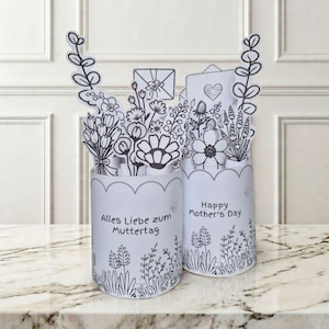 Peut inclure: Deux compositions florales pop-up en papier blanc avec des dessins au trait noirs. L'une dit "Alles Liebe zum Muttertag" et l'autre "Happy Mother's Day". Les compositions ont un bord festonné et des motifs floraux.