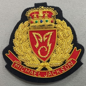 May include: Embroidered patch with a red shield and gold 'M' design, a gold crown, and the name "MICHAEL JACKSON" on a red banner. The patch has a black background and gold laurel leaves.