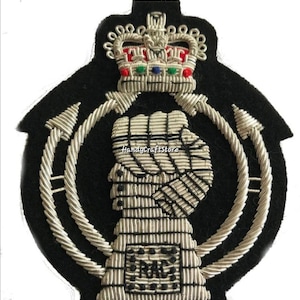 Puede incluir: Parche bordado negro con una corona con detalles rojos, verdes y azules. Presenta un perfil de persona y las letras "RAC" en hilo plateado, rodeadas por un diseño de cuerda plateada.