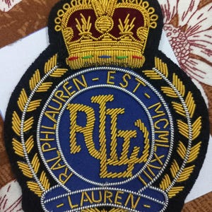 May include: Embroidered patch with a navy blue background, featuring gold-coloured lettering and a crown. The patch reads "RALPH LAUREN" and "EST. MCMXL". The design includes a laurel wreath and intricate detailing.