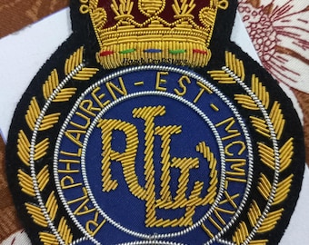 May include: Embroidered patch with a navy blue background, featuring gold-colored lettering and a crown. The patch reads "RALPH LAUREN" and "EST. MCMXL". The design includes a laurel wreath and intricate detailing.