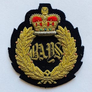 May include: A black embroidered patch featuring a gold laurel wreath and a crown with red and gold accents. The patch has the letters "BAIS" in gold. The patch is likely for a uniform or decorative item.