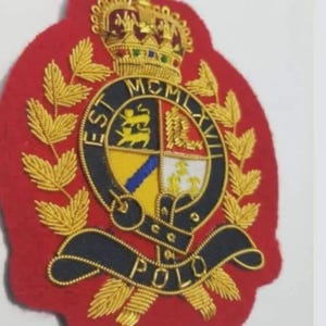 Op de afbeelding: Een rood geborduurd embleem met gouden en zwarte details. Het embleem heeft een kroon, een cirkelvormig embleem met tekst en het woord "POLO" aan de onderkant. Het ontwerp bevat gouden laurierbladeren en ingewikkelde steken.