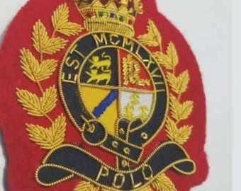 May include: A red embroidered patch with gold and black detailing. The patch features a crown, a circular emblem with text, and the word "POLO" at the bottom. The design includes gold laurel leaves and intricate stitching.