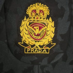 Może przedstawiać: Zbliżenie na ciemnozieloną marynarkę z kwiatowym wzorem. Marynarka posiada haftowaną naszywkę ze złotą koroną, emblematem ptaka i nazwą marki "PRADA" w prostokątnej ramce.