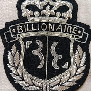Peut inclure: Écusson brodé noir avec une couronne argentée et le mot "BILLIONAIRE" au-dessus d'un motif de bouclier. Le bouclier porte le chiffre "38" et est entouré de feuilles de laurier argentées.