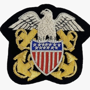 Pode incluir: Patch bordado azul marinho com uma águia prateada, âncoras douradas e um escudo com listras vermelhas e brancas e estrelas azuis. O patch tem uma borda preta e é provavelmente uma insígnia militar.
