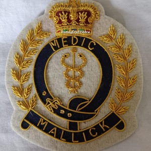 May include: Embroidered patch featuring a gold crown, laurel leaves, and the word "MEDIC" in gold lettering on a navy blue background. The patch also includes the name "MALLICK" and a medical symbol. The patch is on a light gray felt background.