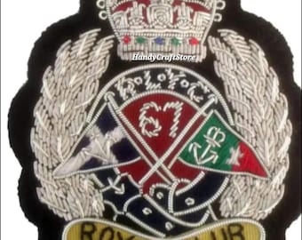 May include: A black embroidered patch with a silver crown, laurel leaves, and crossed flags. The flags are red, green, and blue, with the number "67" and the words "Royal Club" in gold lettering.