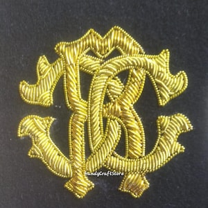 May include: A gold embroidered emblem with a monogram design, set against a black backdrop. The emblem is made with detailed stitching and a decorative border. The text "HandyCraftStore" is visible. The design is likely for clothing or accessories.
