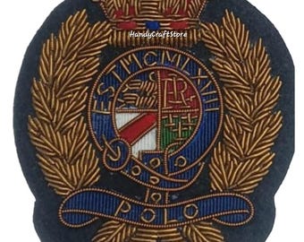 May include: Embroidered patch with a navy blue background. The patch features a gold-colored laurel wreath, a red and gold crown, and a central emblem with text and a Union Jack design. The word "POLO" is embroidered in blue.