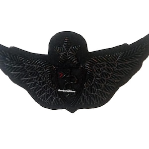 May include: Black embroidered patch in the shape of wings, with a central emblem featuring a star and a medical symbol. The wings have detailed stitching, and the patch has a textured appearance. The text "HandyCraftStore" is visible.
