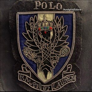 May include: Embroidered patch with the word "POLO" at the top. The patch features a shield design with a crown, tennis racket, and the letters "RL". The words "RALPH LAUREN 67" are at the bottom. The patch is black, blue, silver, and gold.