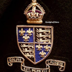 Op de afbeelding: Een goudkleurig draadwapen met een kroon, schild en banners. Het schild is verdeeld in vier secties met blauwe en zwarte achtergronden, met fleur-de-lis en leeuwenmotieven. De banners luiden "RALPH", "LAUREN" en "EST. MCMLXII".