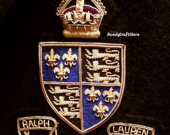 May include: A gold-colored wire crest with a crown, shield, and banners. The shield is divided into four sections with blue and black backgrounds, featuring fleur-de-lis and lion designs. The banners read "RALPH", "LAUREN", and "EST. MCMLXII".