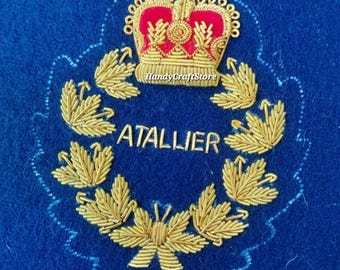 May include: A blue embroidered patch featuring a gold crown with red accents above a gold laurel wreath. The word "ATALLIER" is embroidered in gold within the wreath. The patch has a scalloped edge.