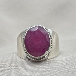 Puede incluir: Anillo de plata con un gran rubí ovalado. El rubí, de color rojo intenso, está engastado en un bisel con un borde tipo cuerda. El anillo tiene una banda ancha, una joya llamativa.