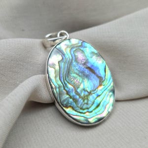 Op de afbeelding: Ovale hanger met een iriserend abalone schelp ontwerp. De schelp heeft wervelende patronen in blauw, groen en paars, gezet in een zilverkleurig metalen frame. De hanger heeft een kleine lus aan de bovenkant voor een ketting.