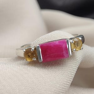 Könnte beinhalten: Ein silberner Ring mit einem großen, rechteckigen, magentafarbenen Edelstein in der Mitte, flankiert von zwei kleineren, runden, gelbbraunen Edelsteinen. Der Ring liegt auf einem neutralen Stoffhintergrund.