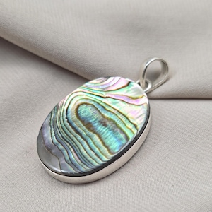 May include: Oval pendant with a polished abalone shell inlay, showcasing iridescent blues, greens, and pinks. The shell is set in a silver bezel with a matching bail for a necklace chain. The pendant rests on a neutral fabric background.