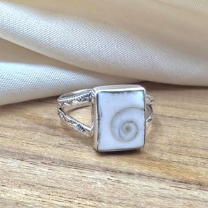 Può includere: Un anello in argento con incastonatura rettangolare e inserto in conchiglia occhio di shiva bianca. La fascia in metallo presenta un design diviso sui lati con motivi geometrici incisi. La conchiglia mostra un motivo a spirale naturale.