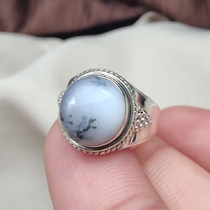 May include: A silver ring with a large, round, pale blue gemstone with dark, tree-like inclusions. The ring has a detailed, textured band and a decorative setting around the stone, showcasing a unique, natural design.