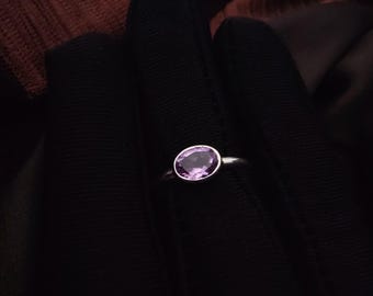 May include: A silver ring featuring a large, oval-cut amethyst gemstone. The ring's band is thin and polished, and the gemstone is a vibrant shade of purple. The ring is set against a dark background, highlighting its elegant design.